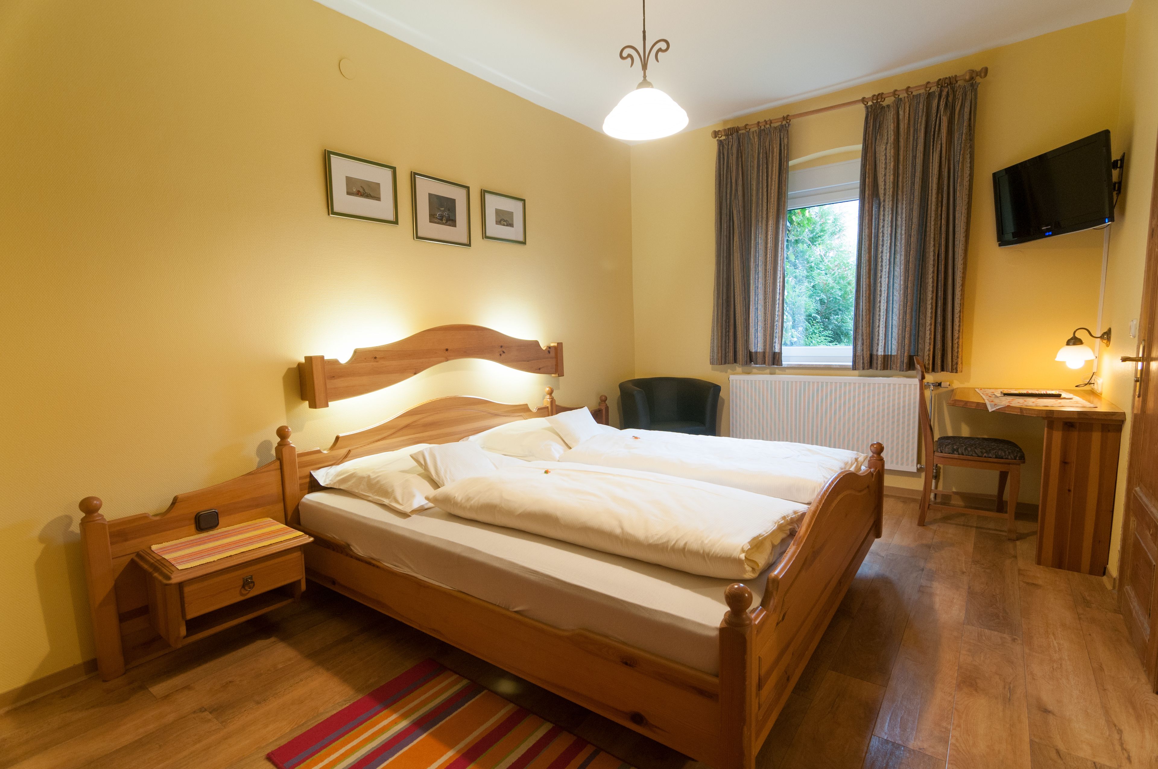 Una cama de madera con una mesita de noche, un escritorio con una silla, un televisor en la pared y una ventana con cortinas.