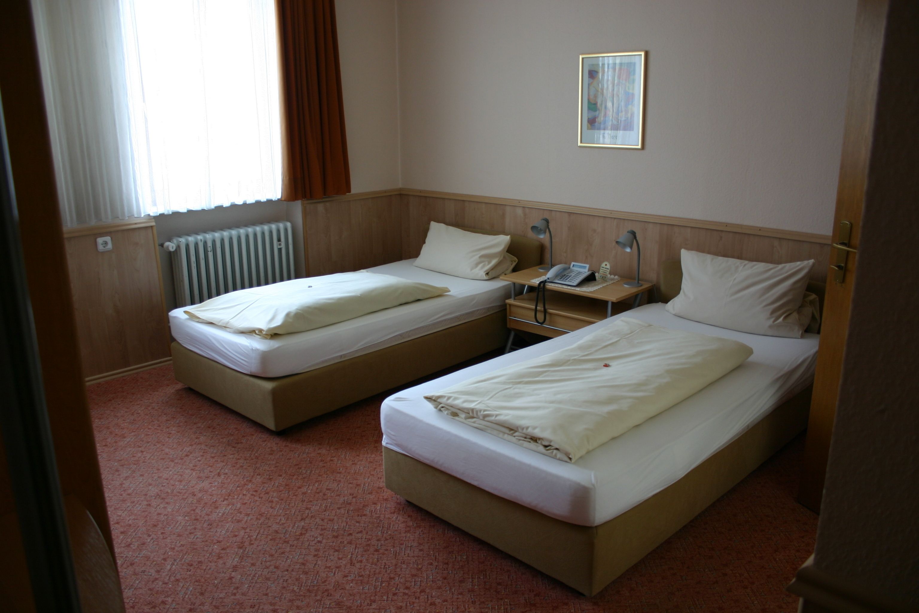 Habitación de hotel con dos camas individuales, una mesita de noche con lámpara, un radiador y una ventana con cortinas.