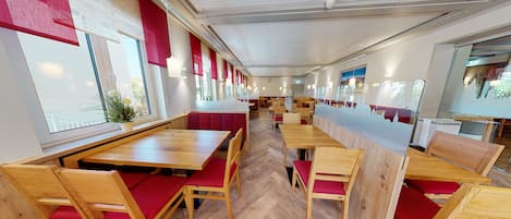 Een modern restaurantinterieur met houten tafels en rode fauteuils, grote ramen en een visgraatvloer.