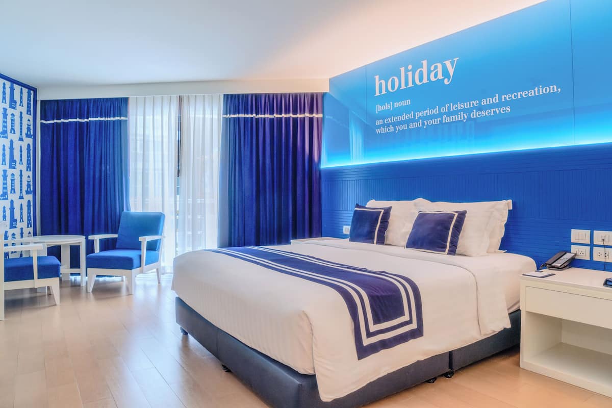 Una habitación de hotel con una cama grande, un escritorio, dos sillas y un televisor montado en la pared que muestra la palabra 'holiday'.