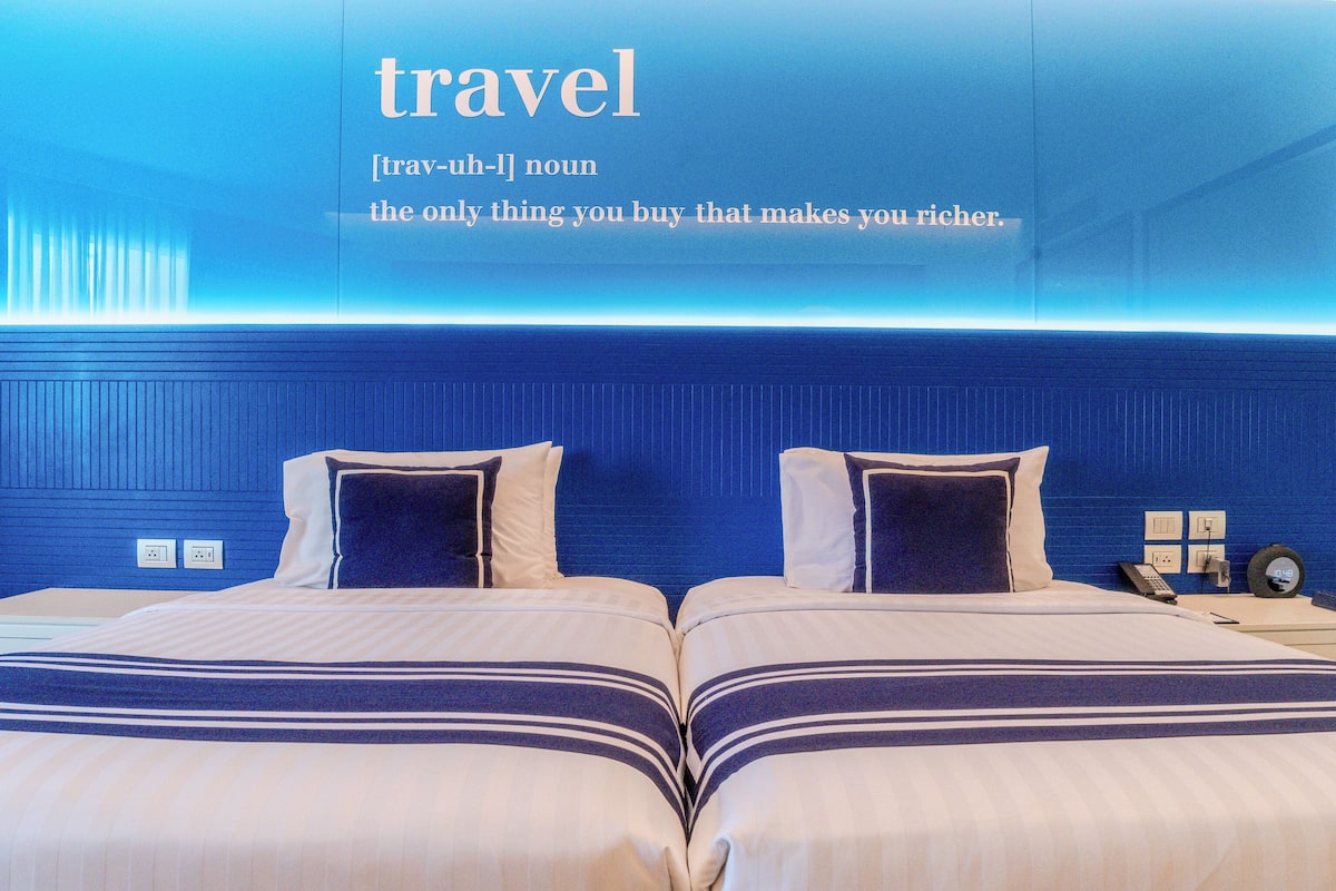 Habitación de hotel con dos camas, una pared azul y una mesita de noche con un teléfono y un reloj.
