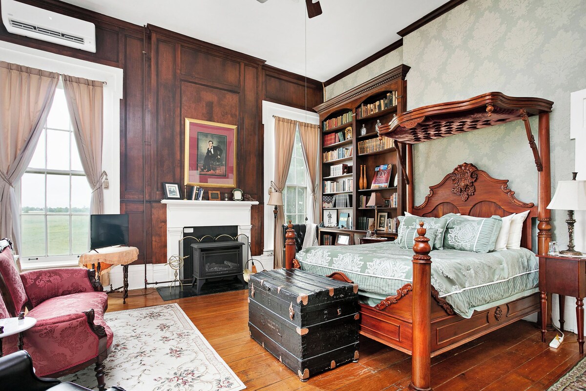 A traditional bedroom with a four-poster bed, a fireplace, a bookshelf, and a vintage suitcase.