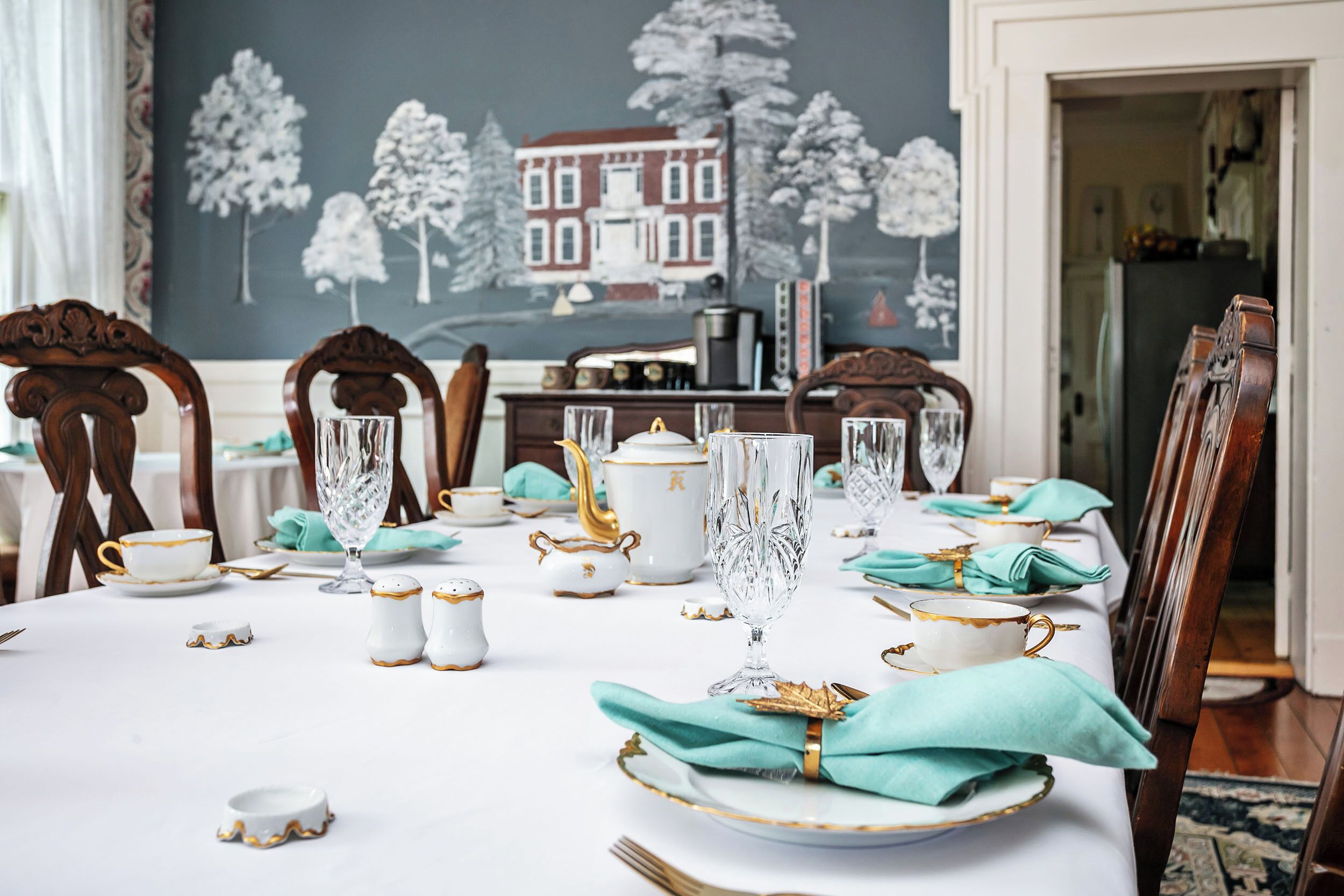 A dining table set with white and gold china, crystal glasses, and teal napkins.