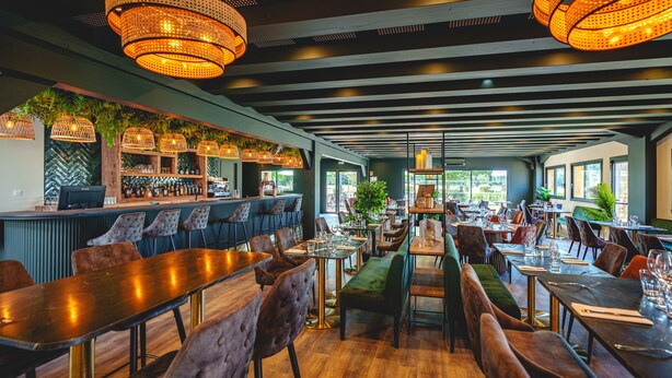 Un restaurante con mesas y sillas de madera, una zona de bar y lámparas colgantes.