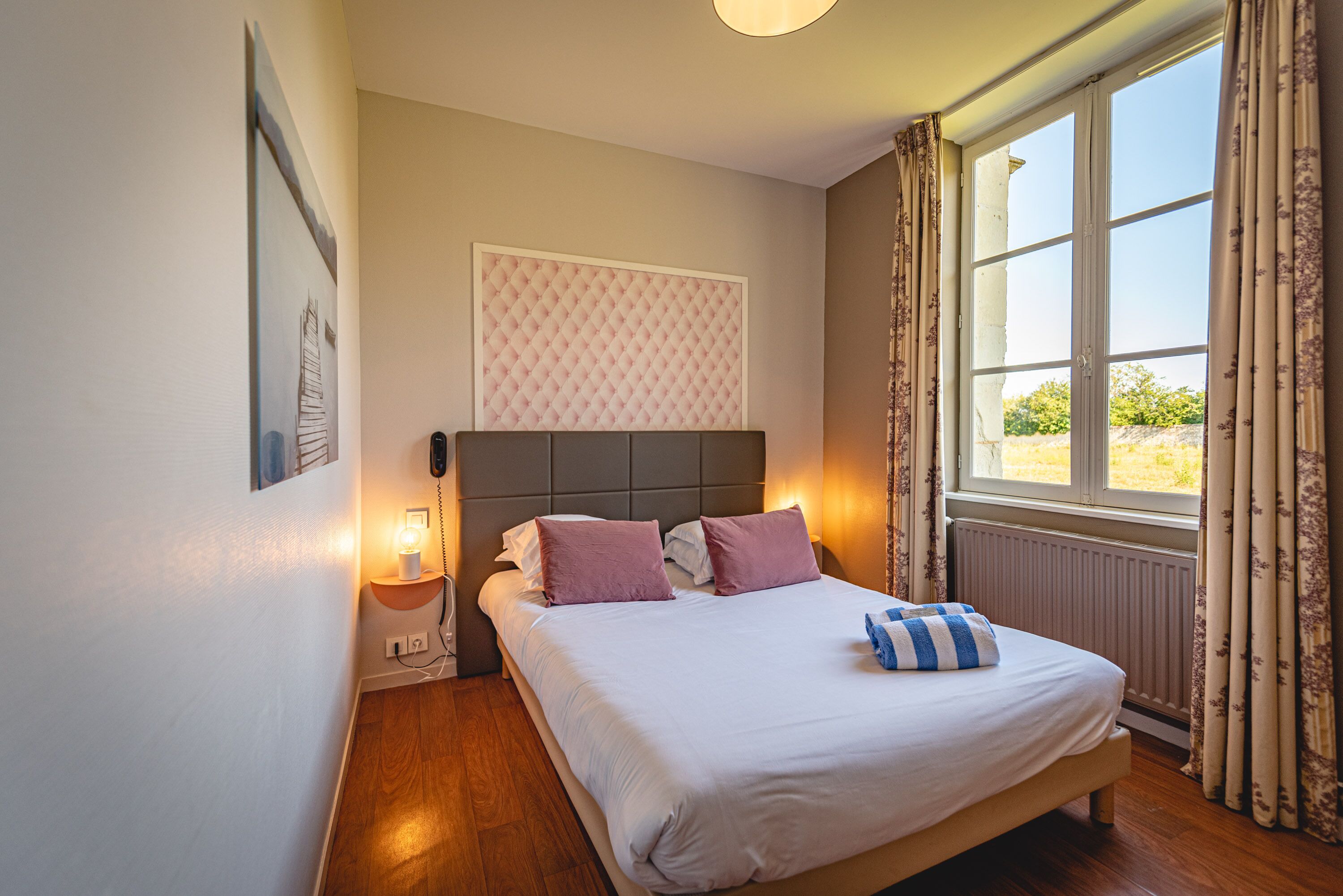A bedroom with a bed, a headboard, a bedside table with a lamp, a window with curtains, and a wall-mounted telephone.
