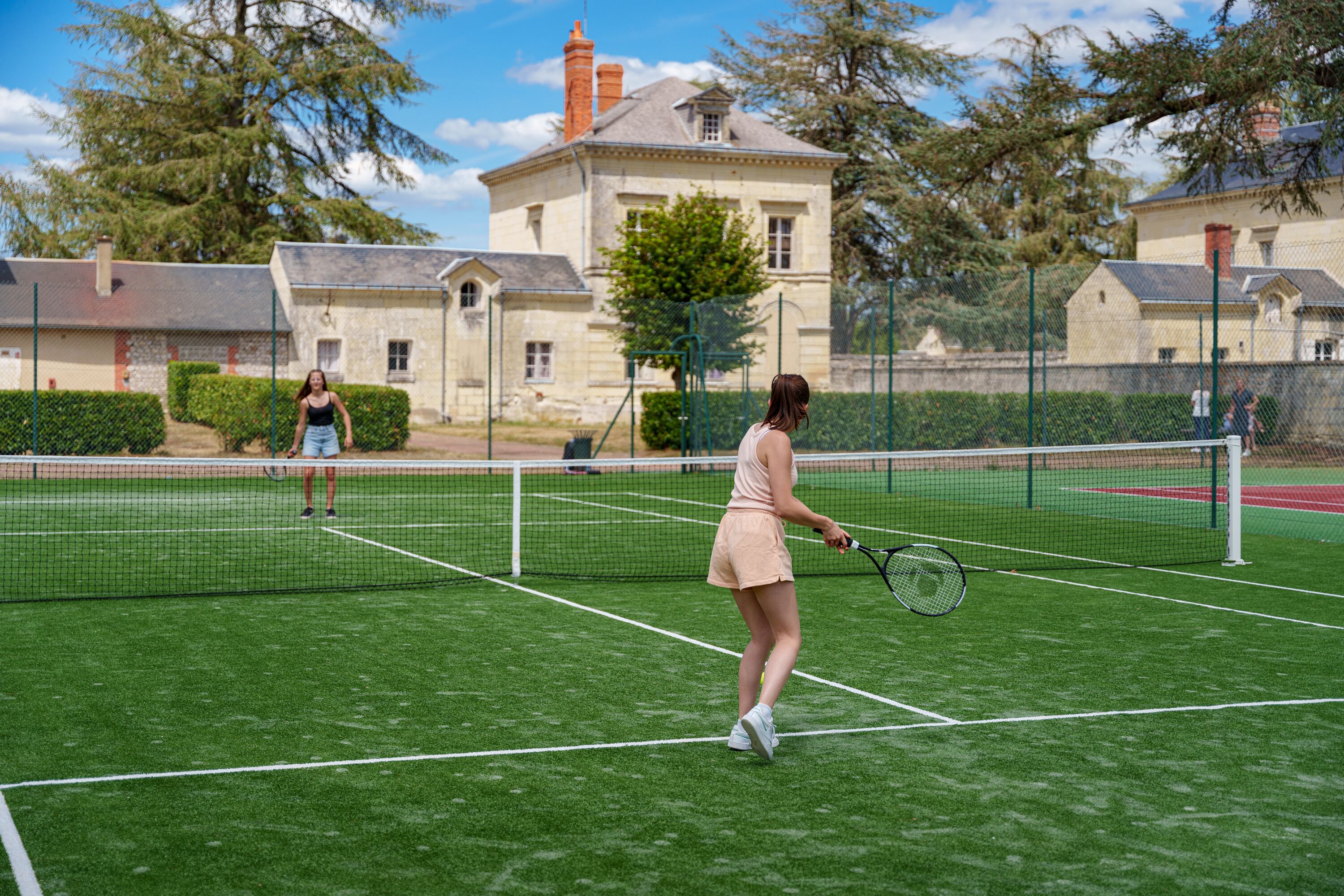 A tennis court with two players in action, surrounded by a fence and a large building in the background.