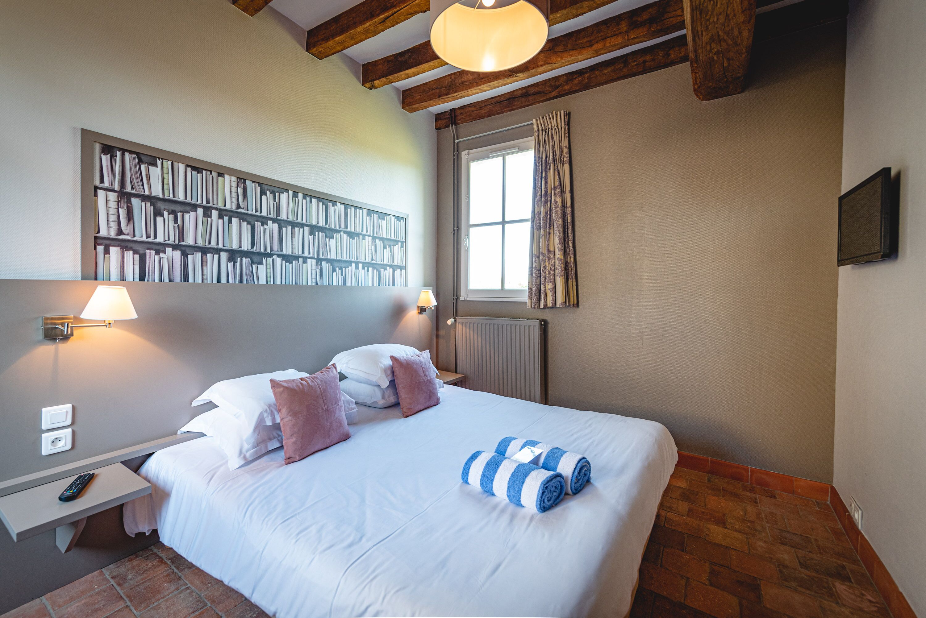 Ein ordentlich bezogenes Bett mit weißen Leinen und blau gestreiften Handtüchern, ein Bücherregal mit Büchern, ein Fenster mit Vorhängen und ein Holzbalken an der Decke.