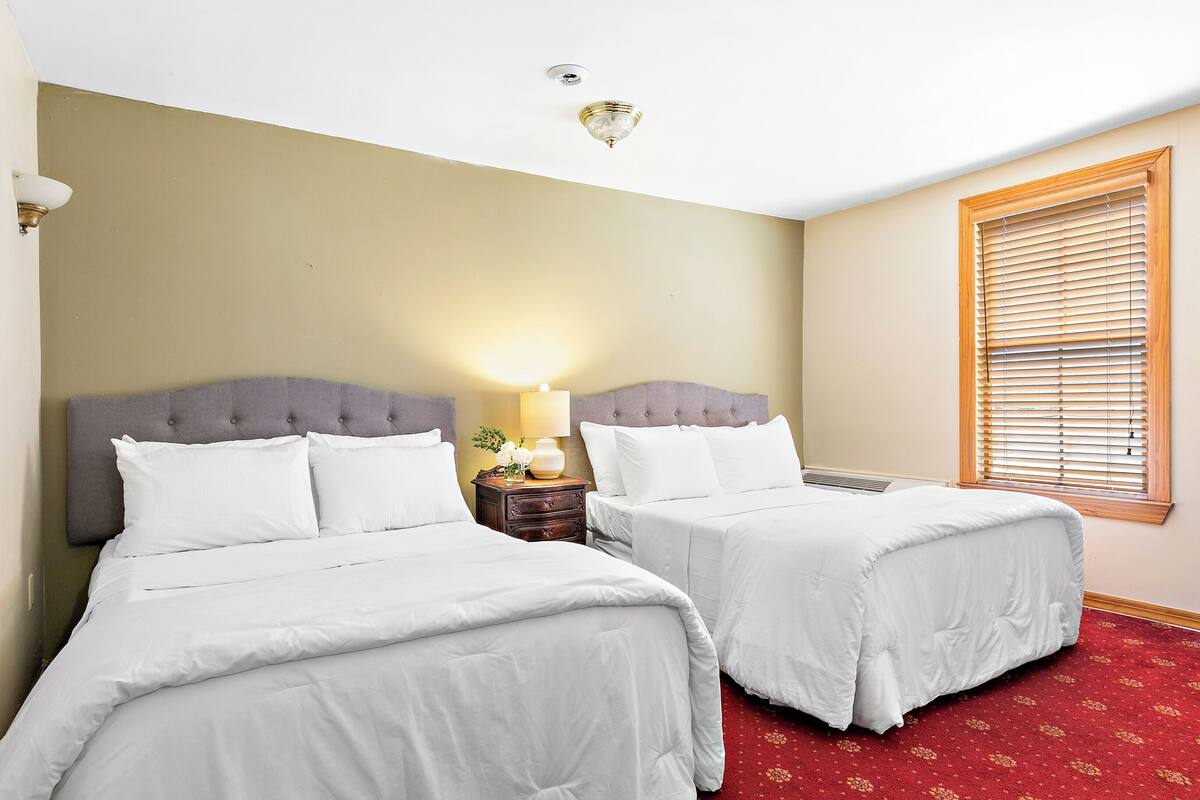 A hotel room with two beds, a nightstand with a lamp, a window with blinds, and a red carpet.