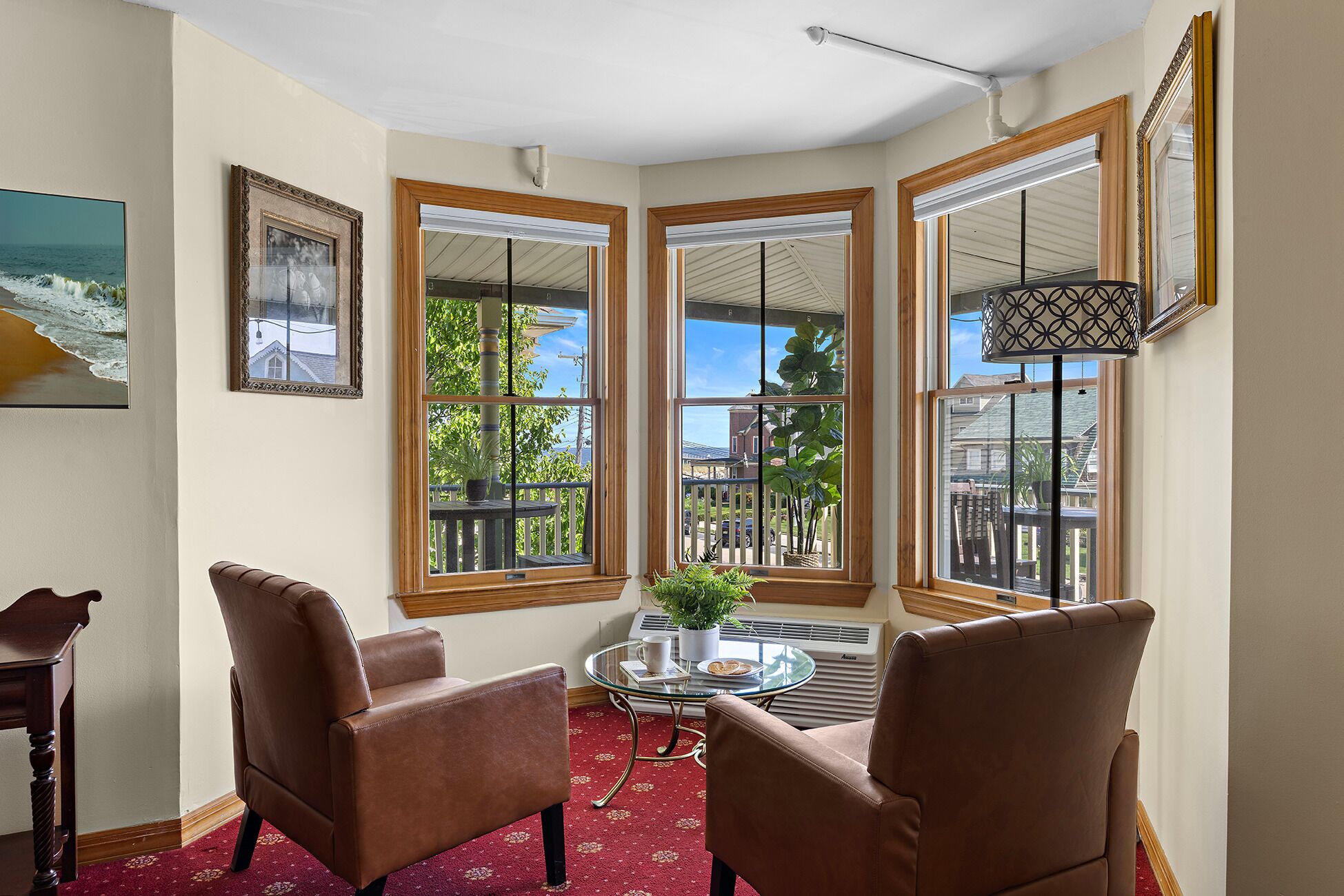 A corner room with three large windows, a round glass table with chairs, a potted plant, and a framed picture on the wall.