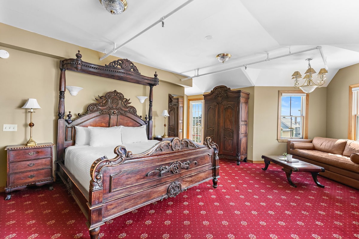 A bedroom with a large ornate bed, a wooden wardrobe, a leather sofa, and a red patterned carpet.