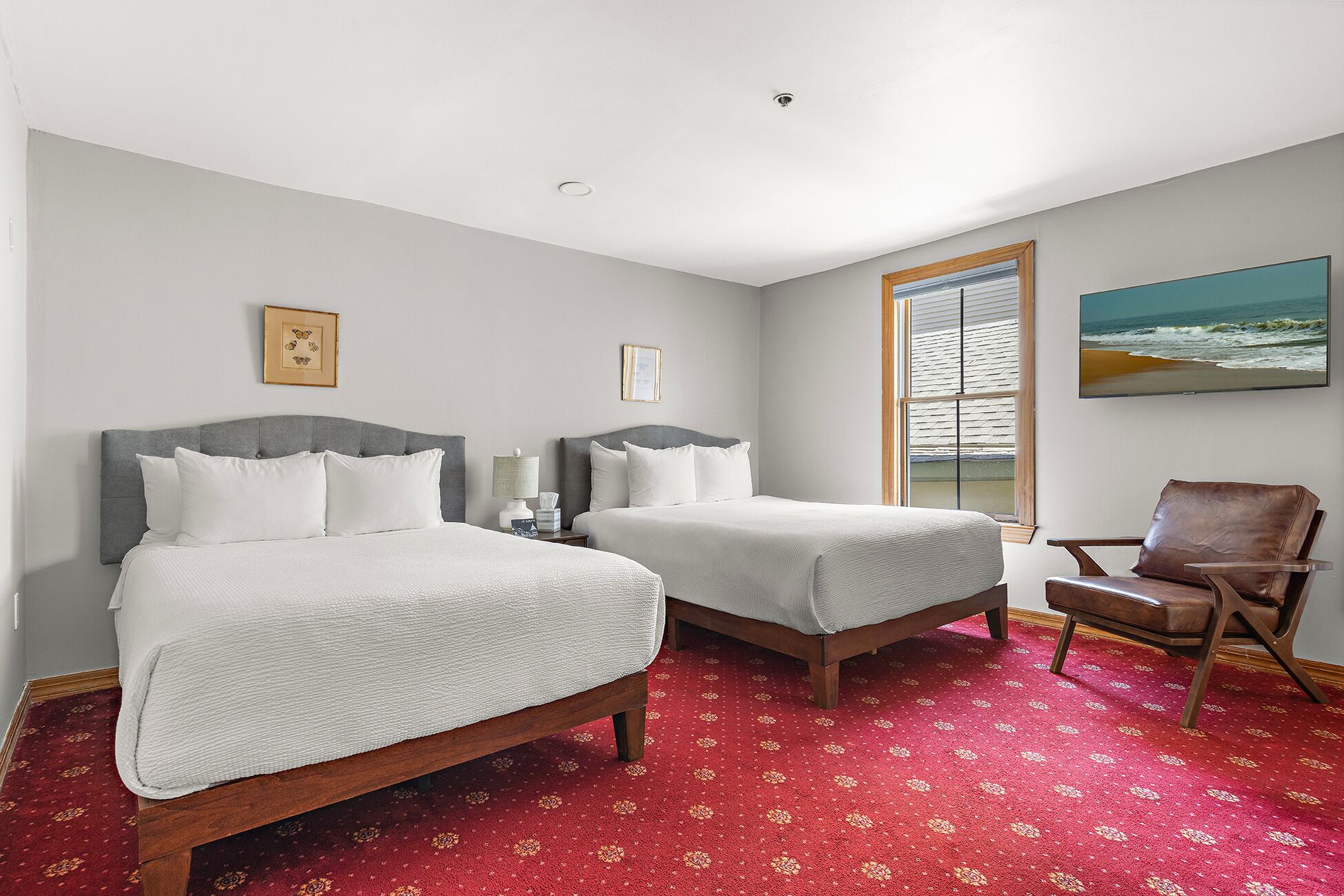 A hotel room with two beds, a red patterned carpet, a wooden chair, and a beach view through the window.