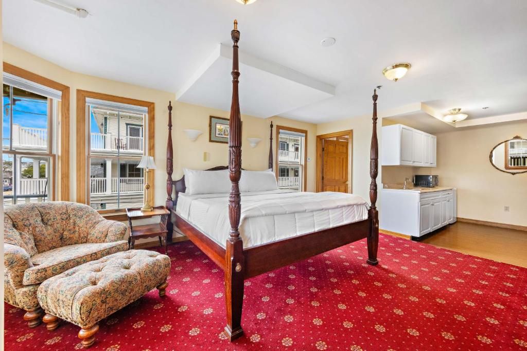 A four-poster bed with a red patterned carpet, a wooden nightstand, a floral armchair, and a white dresser in a bedroom with large windows and a balcony view.