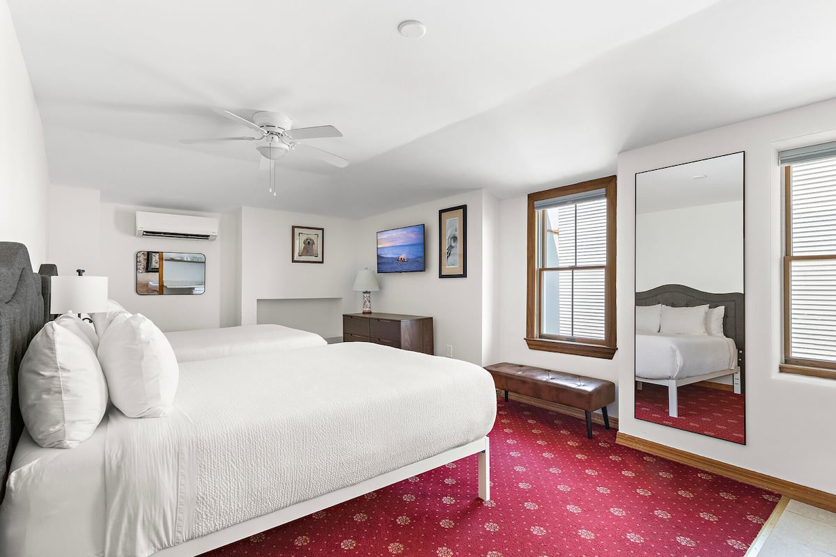 A bedroom with a bed, a ceiling fan, a TV, a fireplace, a bench, and a large mirror.