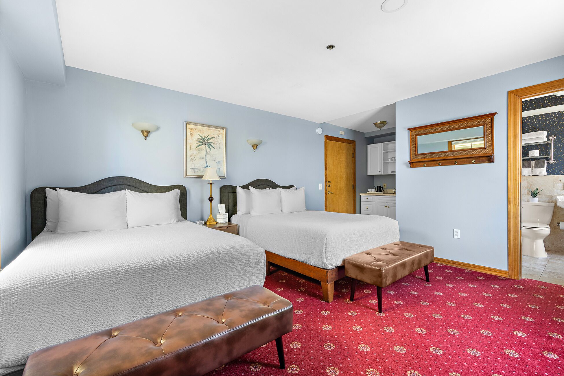 Habitación de hotel con dos camas, alfombra roja con diseño, banca de madera, un cuadro, un espejo y una puerta que da a un baño.