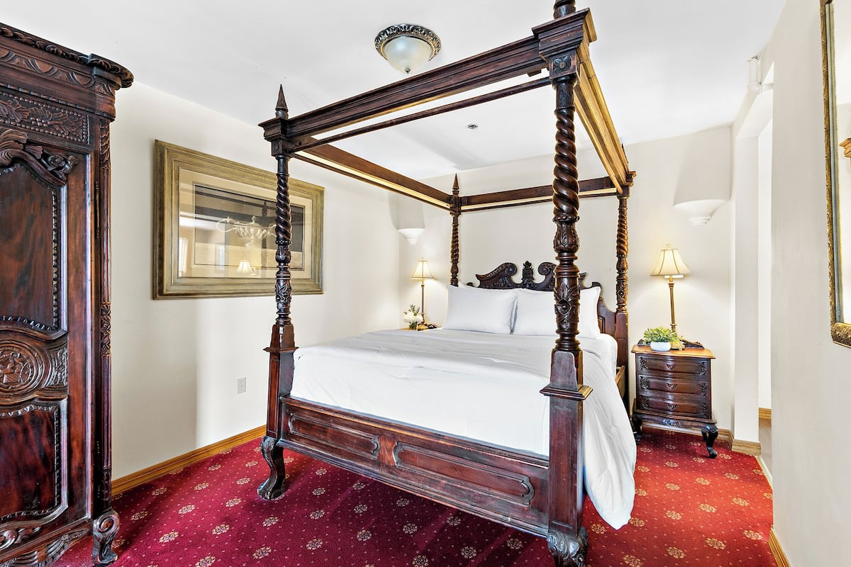 A four-poster bed with a red carpet, a wooden nightstand with a lamp, and a framed picture on the wall.