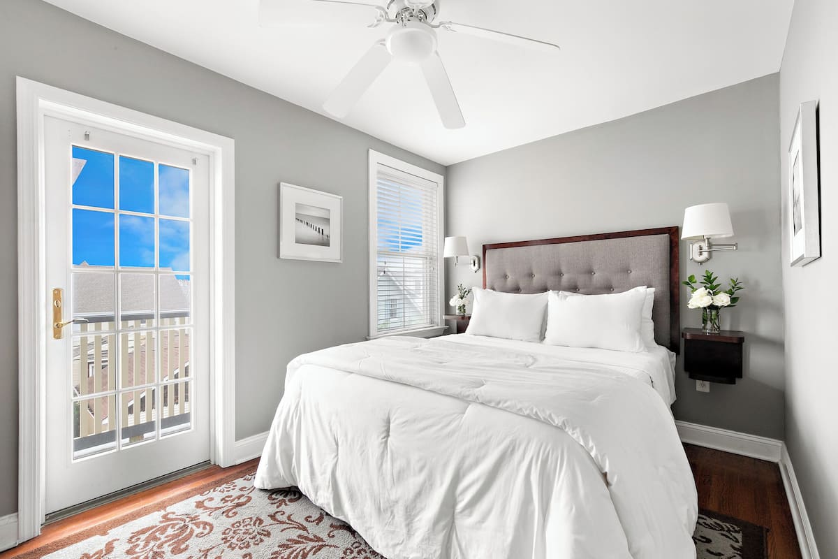 A bedroom with a large bed, a ceiling fan, a window with blinds, and a door with a view of the outside.