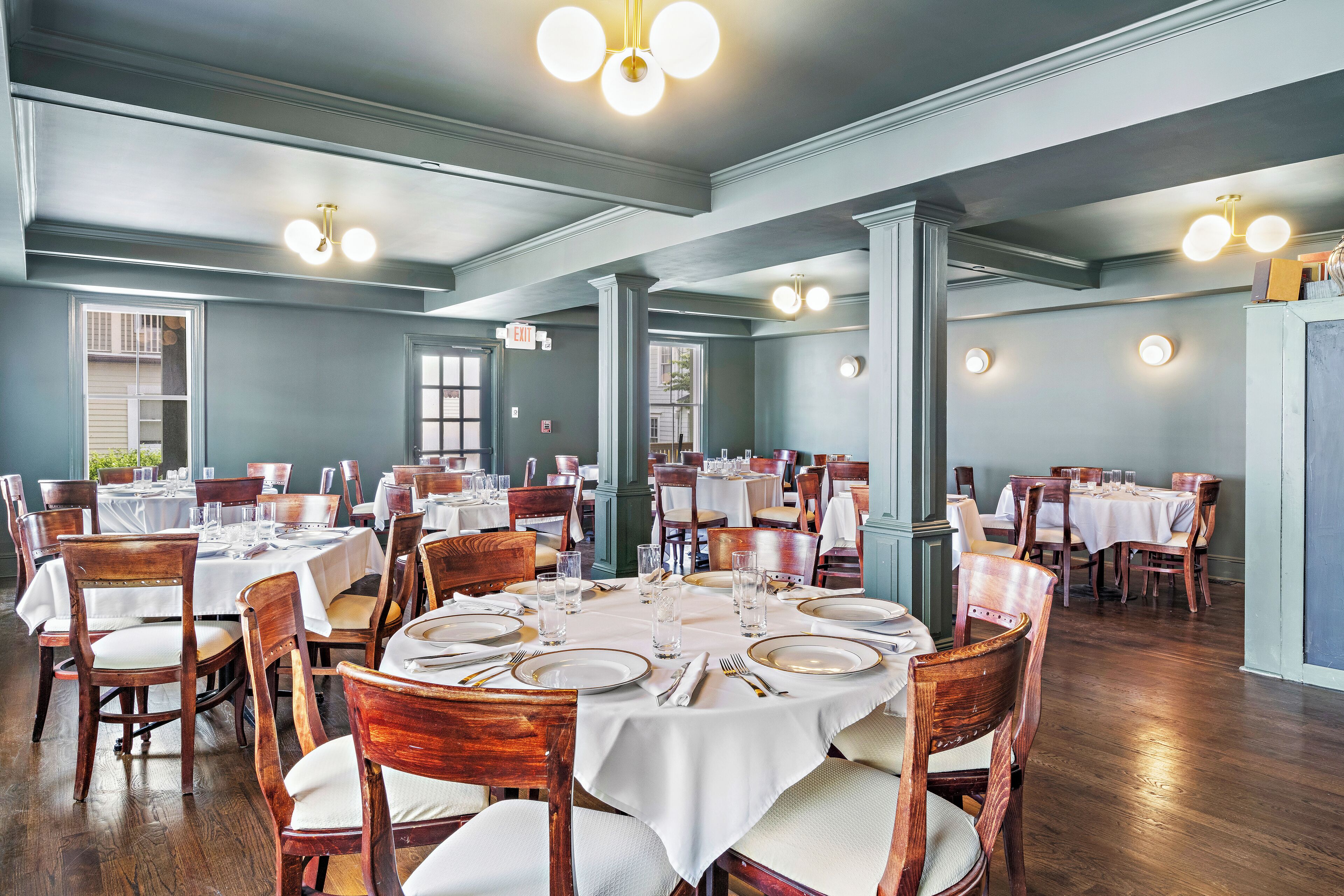 A dining area with round tables set for four, wooden chairs, and white tablecloths.