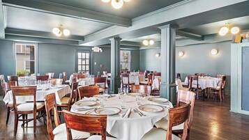 A dining area with round tables set for four, wooden chairs, and white tablecloths.