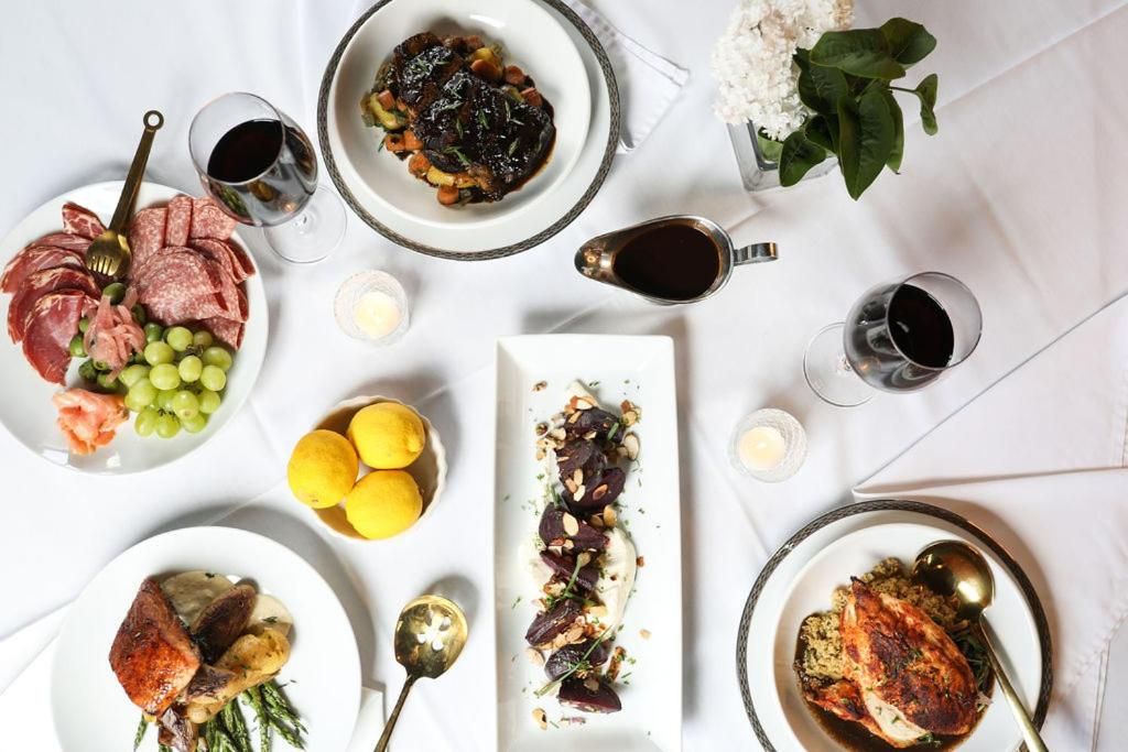 A table set with various dishes, including meats, fruits, and vegetables, accompanied by glasses of red wine.