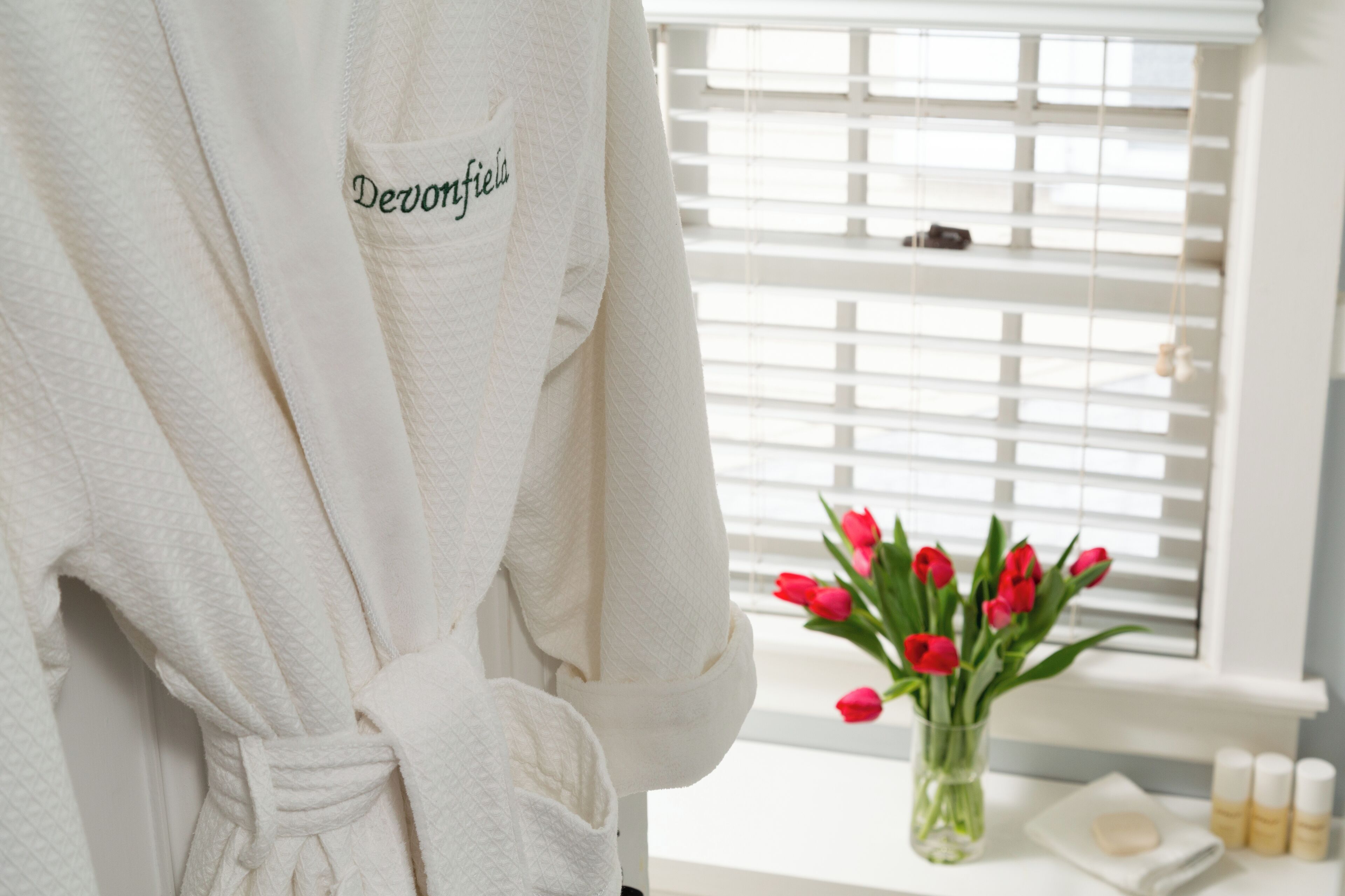 A white robe with "Devonfield" embroidered on it, a window with white blinds, and a vase with red tulips on a table.