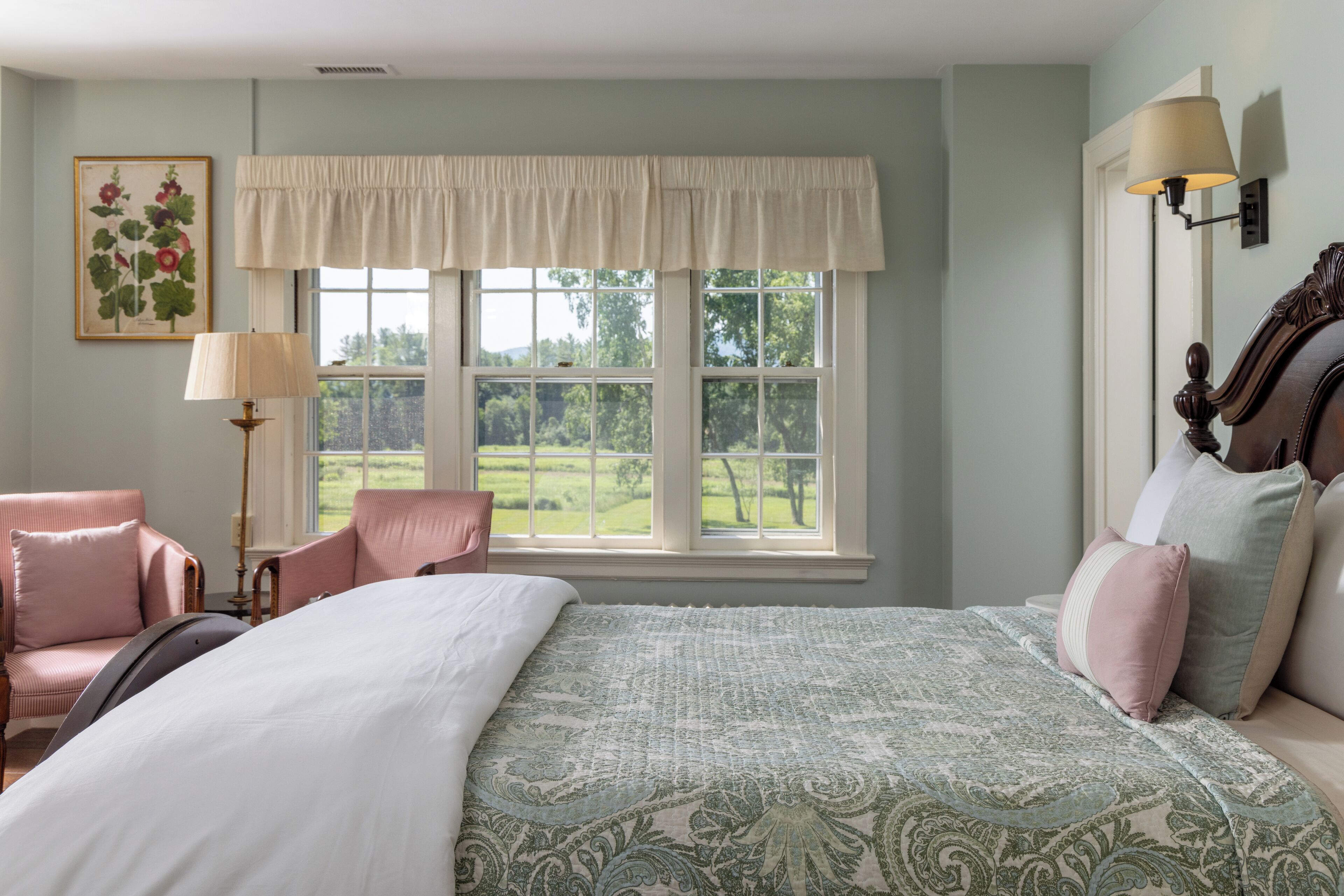 A bedroom with a large bed, a window with a view of a green landscape, a chair, and a lamp.