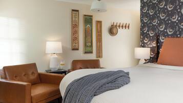 A hotel room with a bed, two chairs, a lamp, and decorative wall art.
