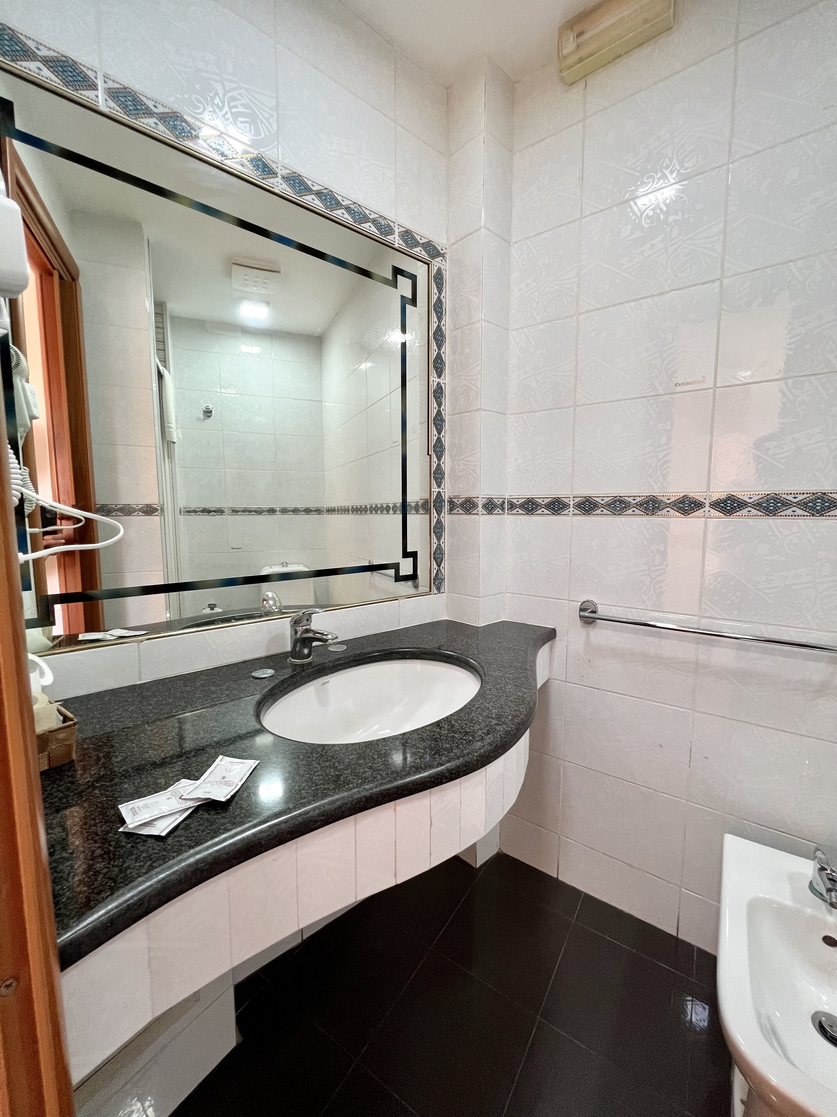A bathroom with a black granite sink, a mirror with a decorative frame, white tiled walls, and a black floor.