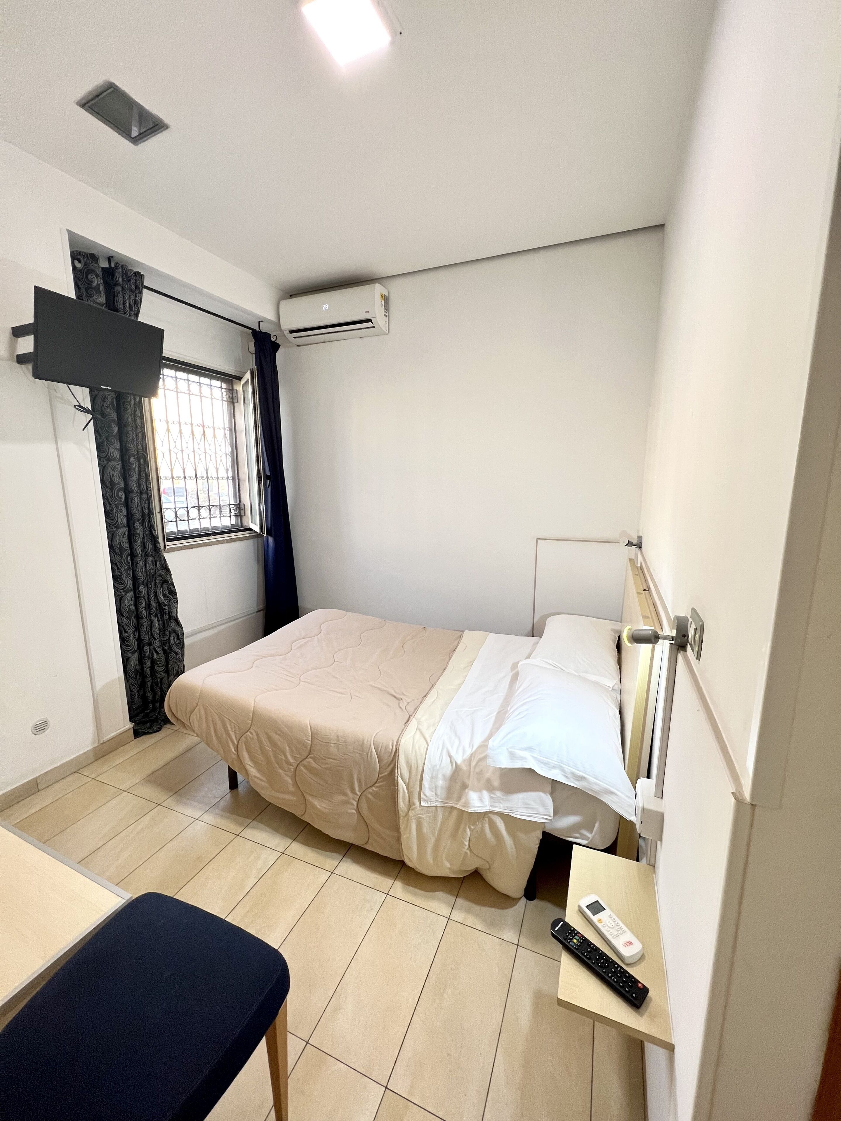 A small, clean room with a bed, a desk, a chair, a window with curtains, an air conditioning unit, and a remote control on the desk.