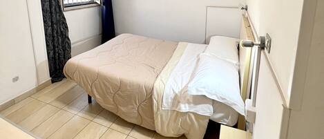 A small, clean room with a bed, a desk, a chair, a window with curtains, an air conditioning unit, and a remote control on the desk.