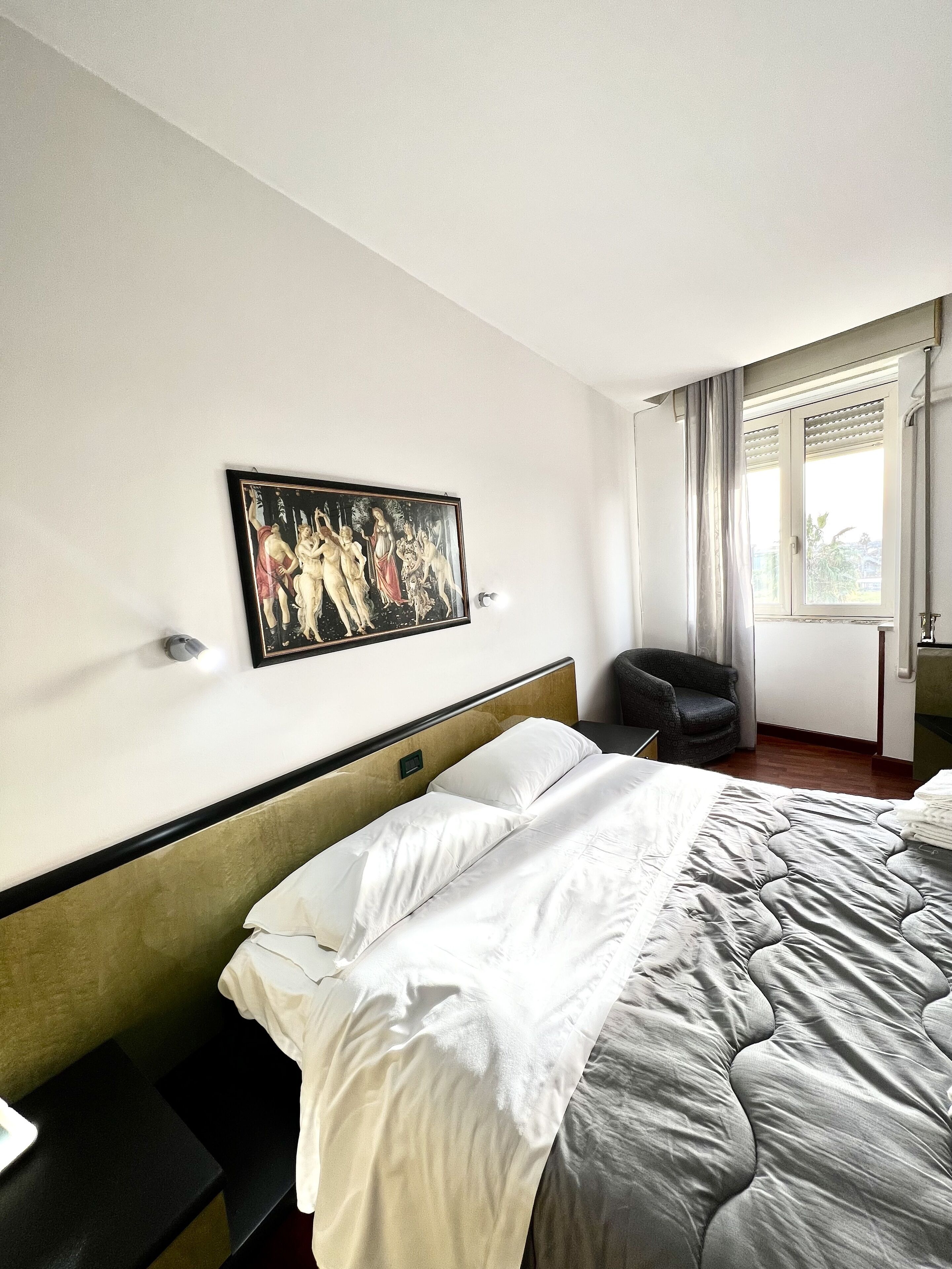 Habitación de hotel con una cama grande, un cuadro en la pared, una silla y un ventanal con cortinas.