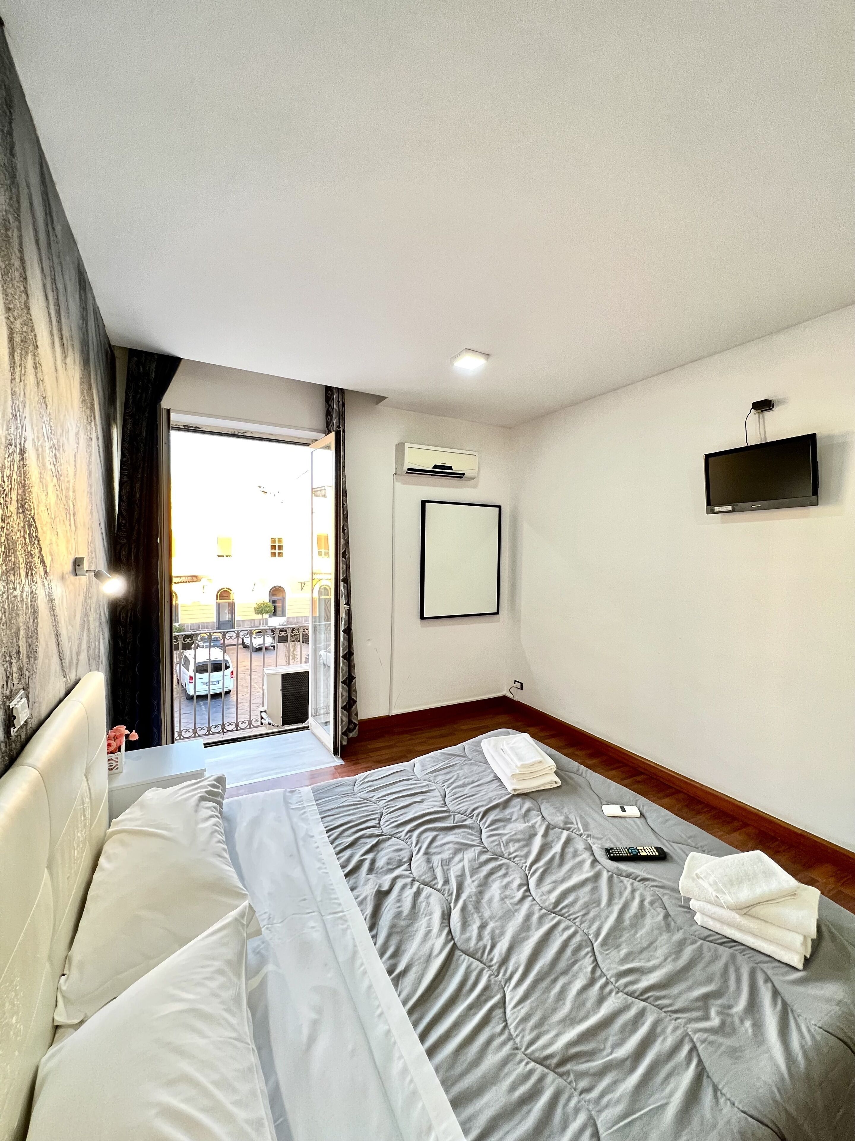 A hotel room with a bed, a flat-screen TV mounted on the wall, an air conditioning unit, and a view of the outside through a glass door.