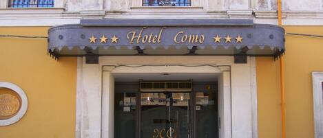 Entrada de un hotel con un cartel que dice "Hotel Como" y tres banderas.