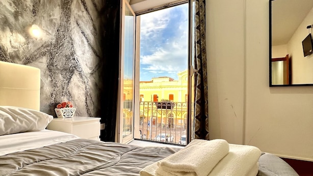 A hotel room with a large bed, a window with a view of buildings, a bedside table with a lamp, and a mirror on the wall. - Hotel Como (Syracuse)