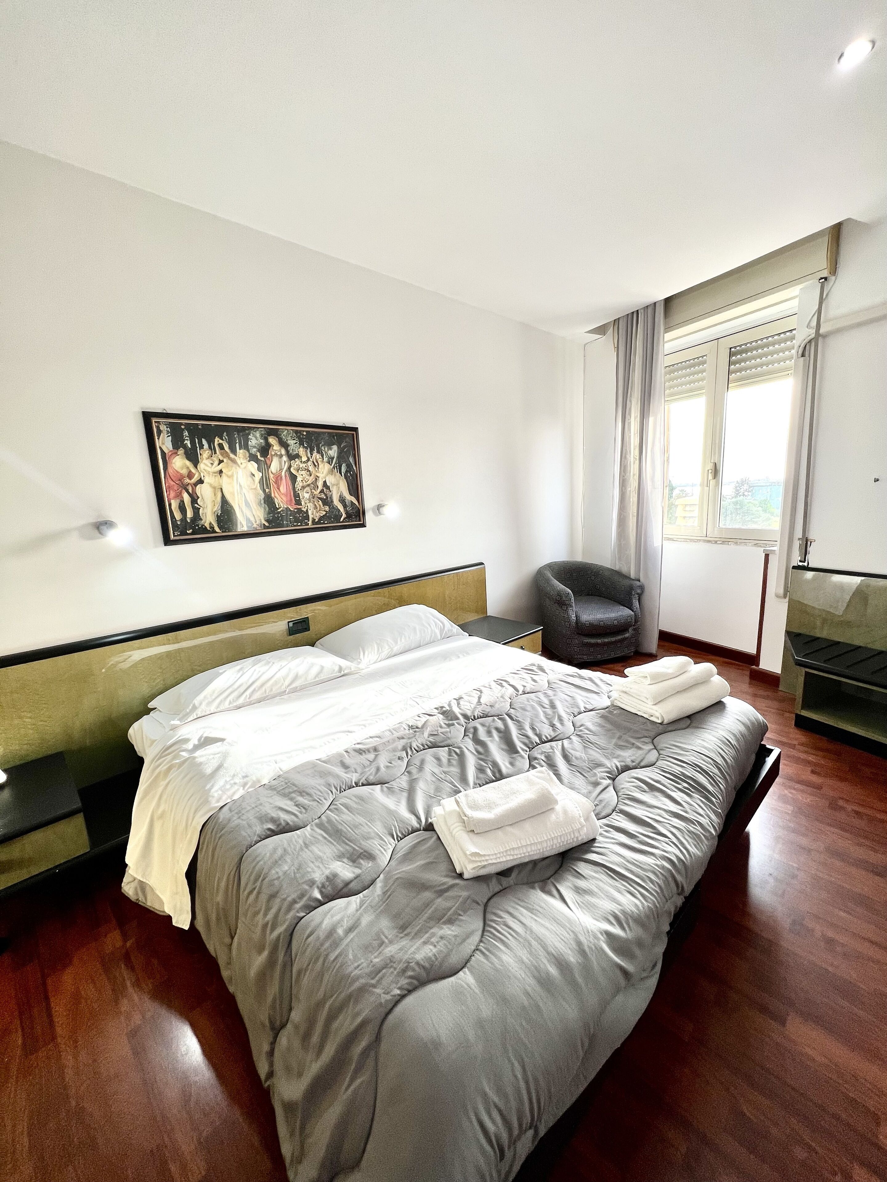 Quarto de hotel com uma cama grande, um quadro na parede, uma cadeira e uma janela com cortinas.