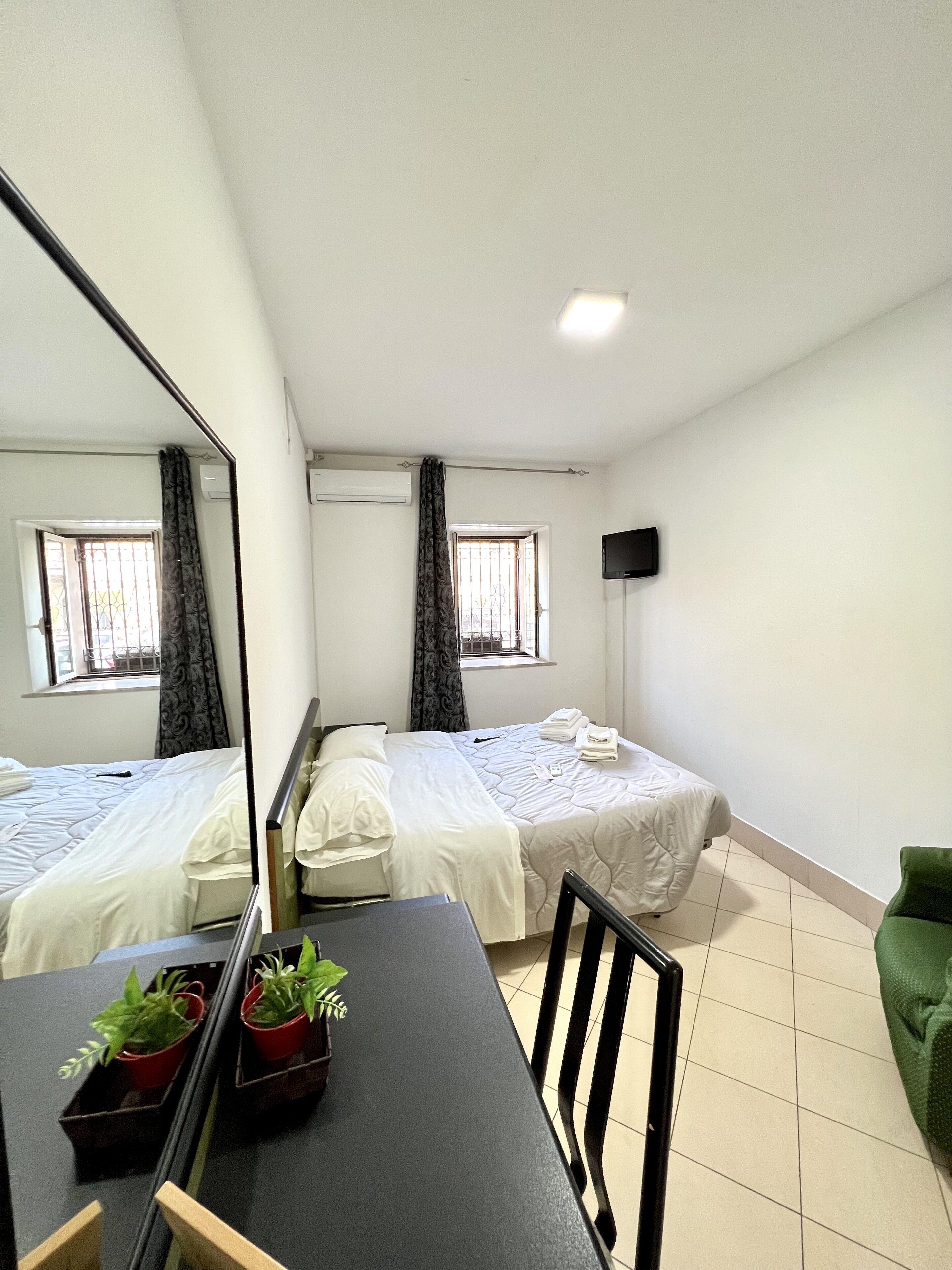 Quarto de hotel com duas camas, uma mesa de jantar, plantas em vasos, uma cadeira verde e uma TV na parede.