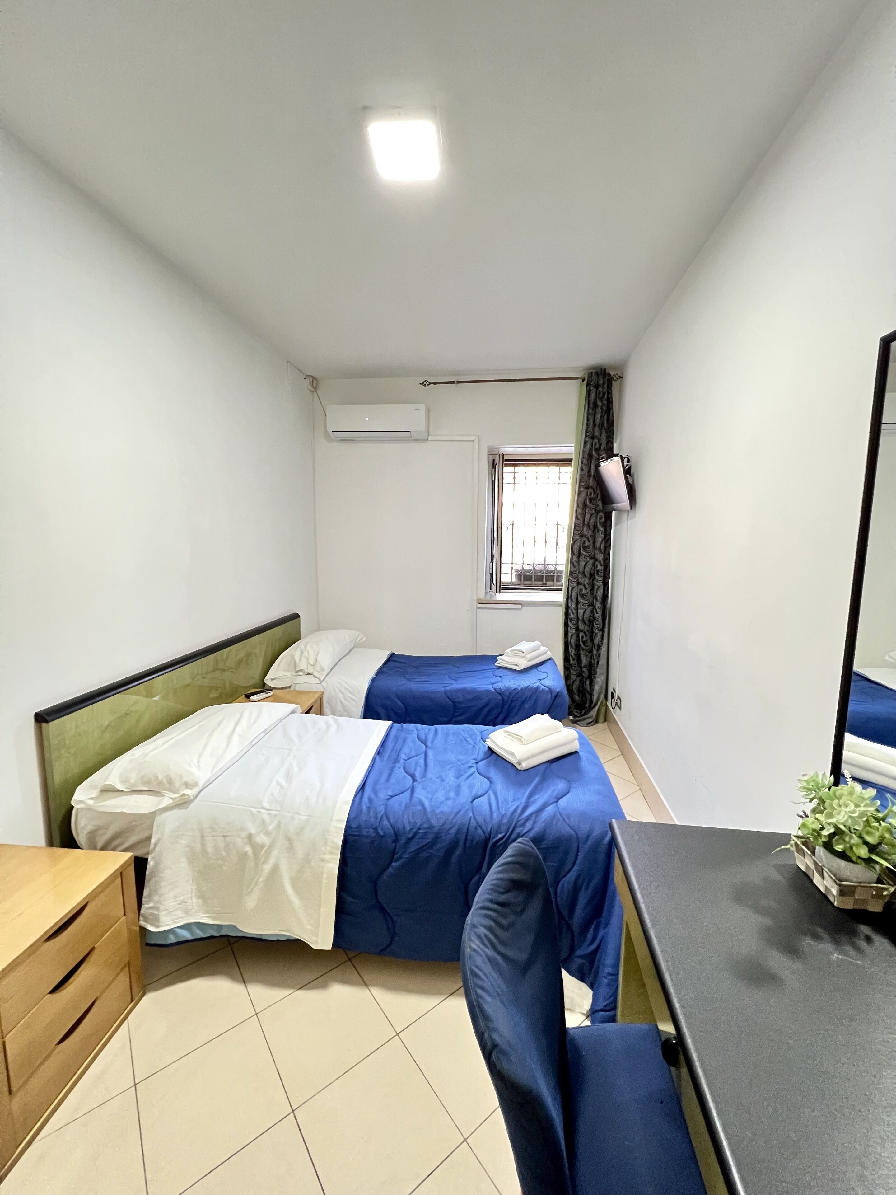 Una habitación pequeña y limpia con dos camas, un escritorio, una silla, un espejo y una ventana con cortinas.