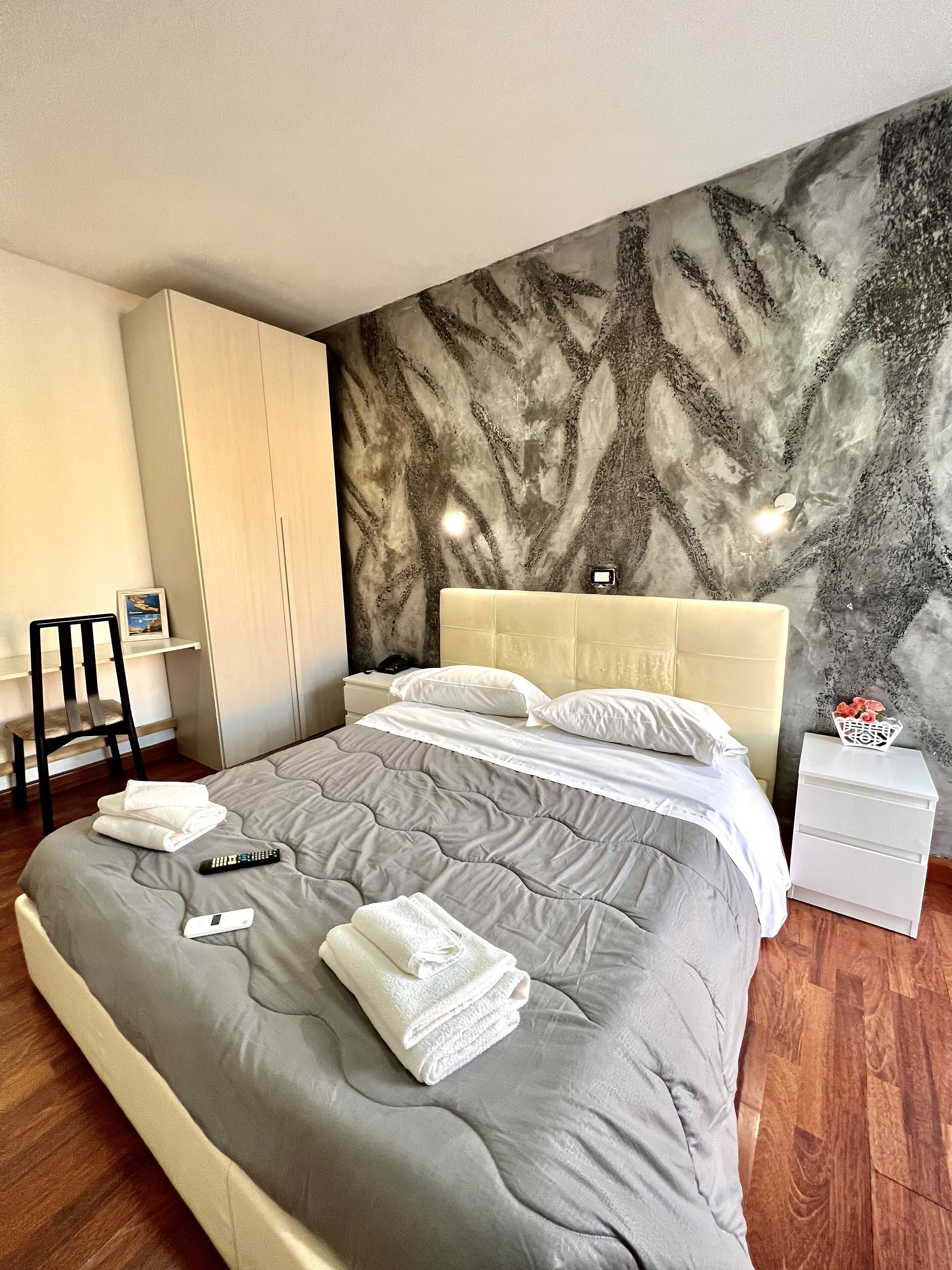 A neatly made bed with a grey quilt, white pillows, and folded towels. A remote control and a thermostat are on the bed. There is a white nightstand with a vase of flowers. The room has a wooden floor and a wall with a textured, abstract design.