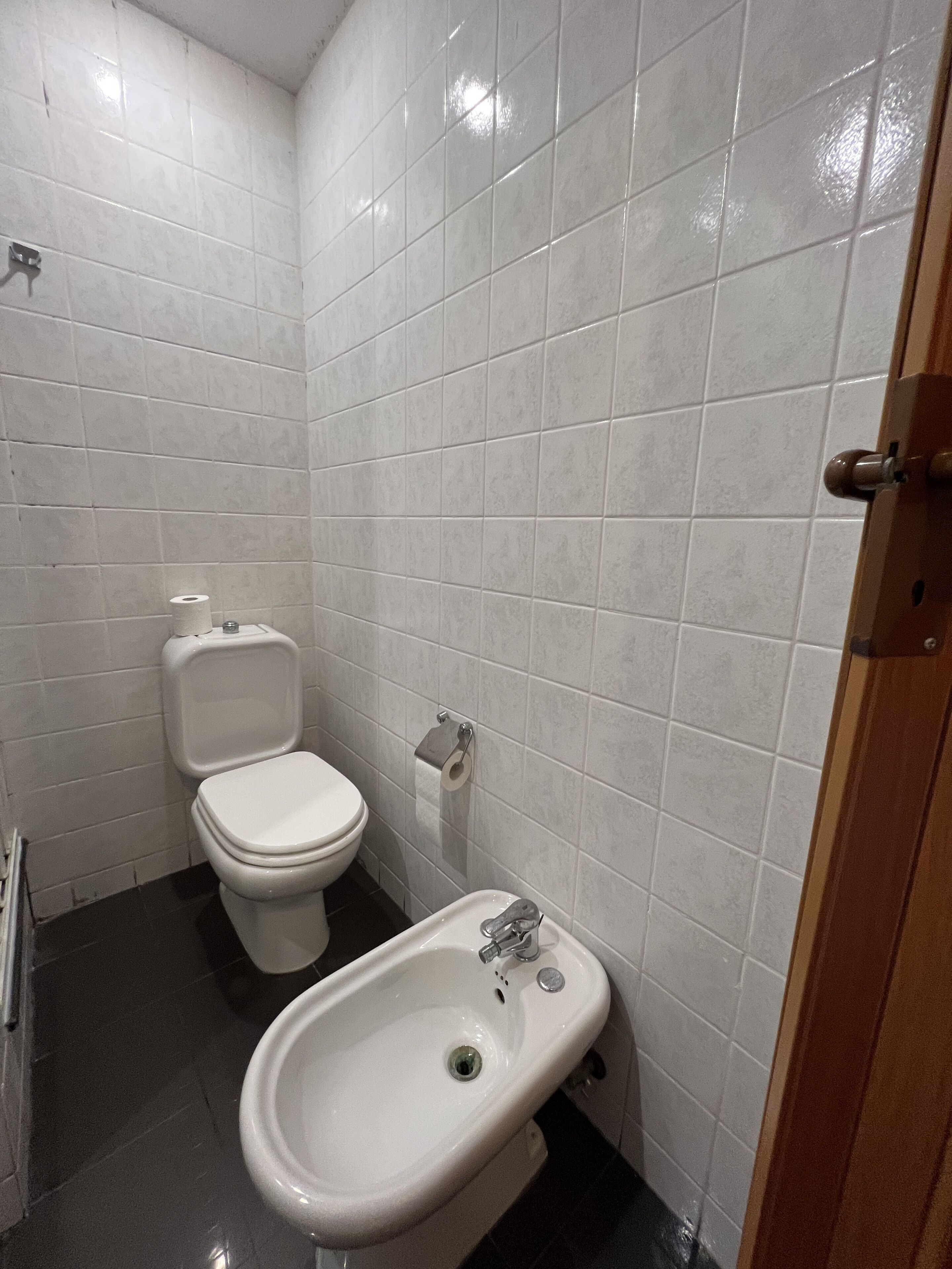 Een kleine badkamer met witte tegels, een toilet en een bidet.
