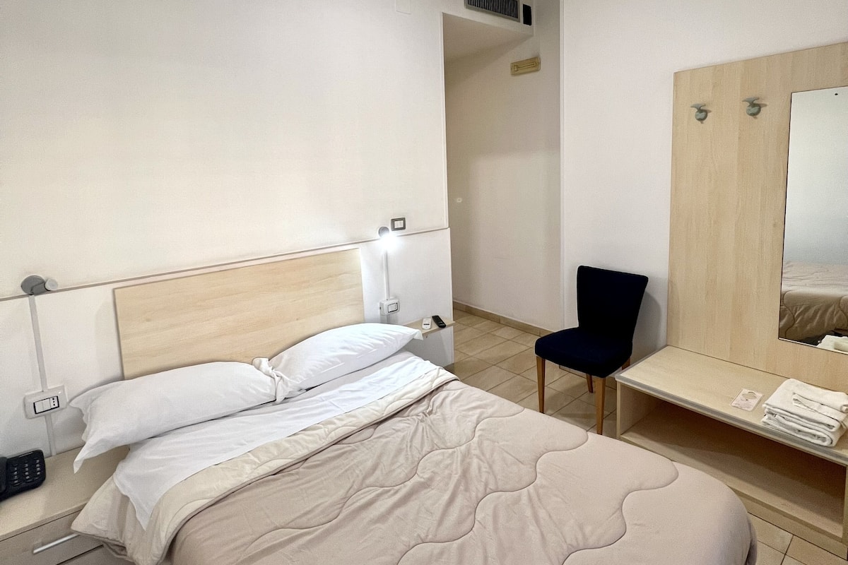 A modern hotel room with a bed, bedside table, chair, and a mirror.
