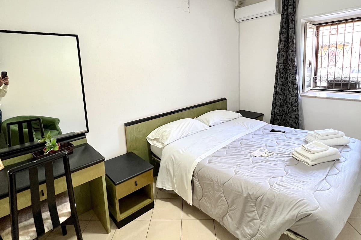 A bedroom with a bed, a desk, a chair, a mirror, and a window with curtains.