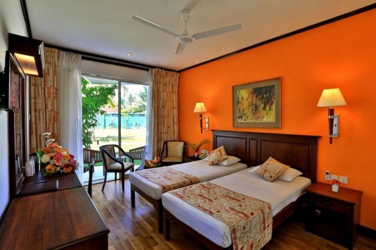 A hotel room with two beds, a desk, a chair, a ceiling fan, and a view of the outdoors.