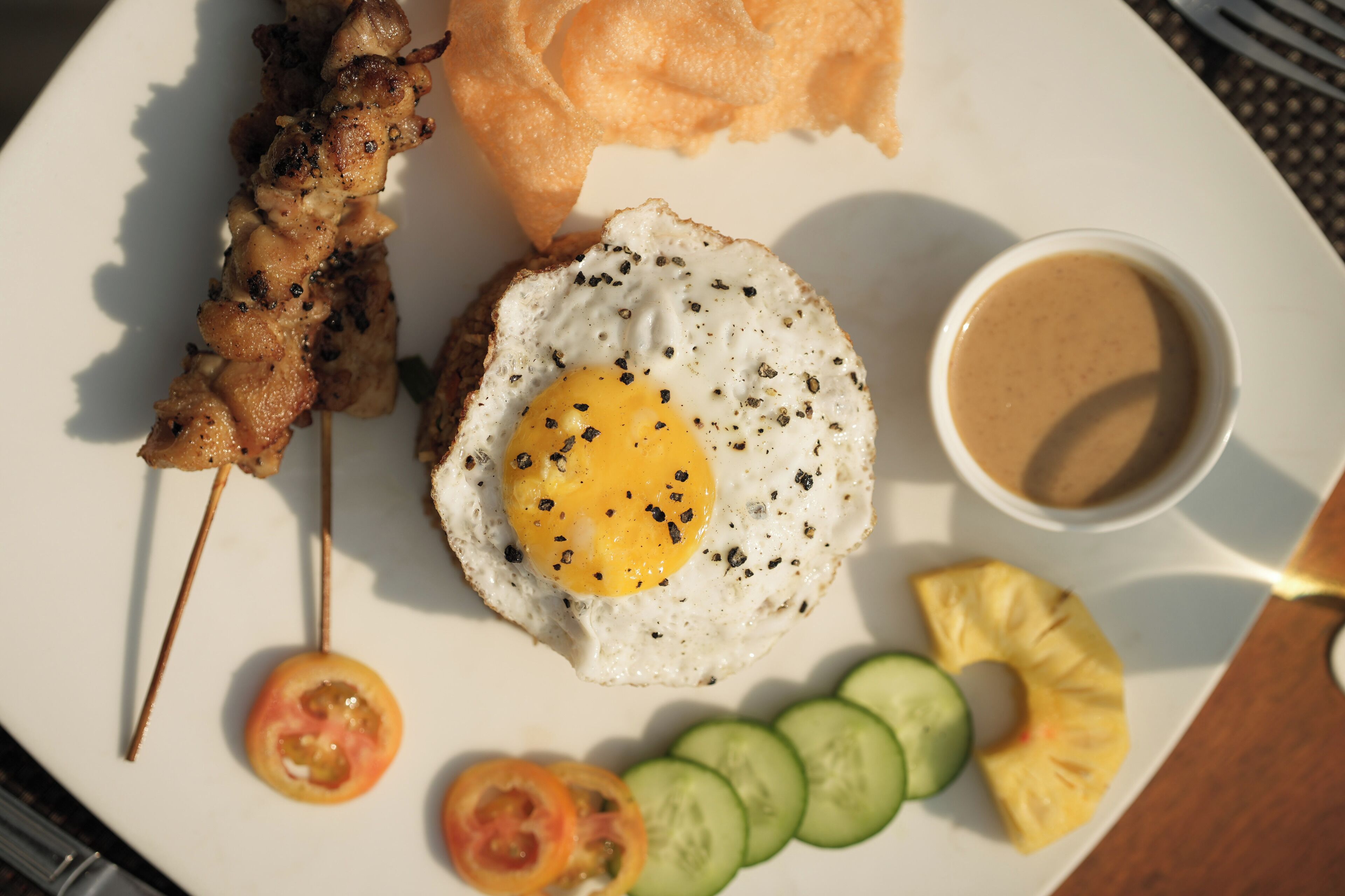 A plate with a fried egg, a skewer of meat, a slice of bread, a small bowl of sauce, and sliced vegetables.
