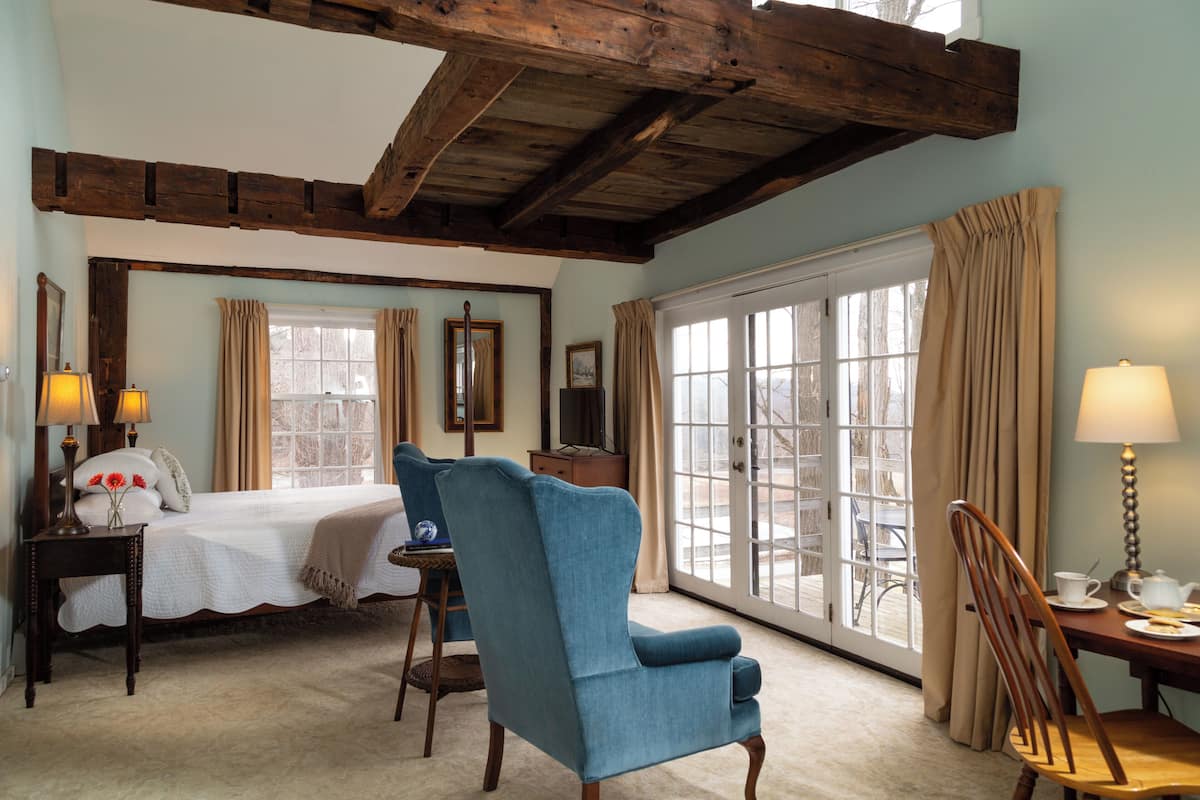 A bedroom with a large bed, a blue armchair, a wooden desk, and a view through a glass door.