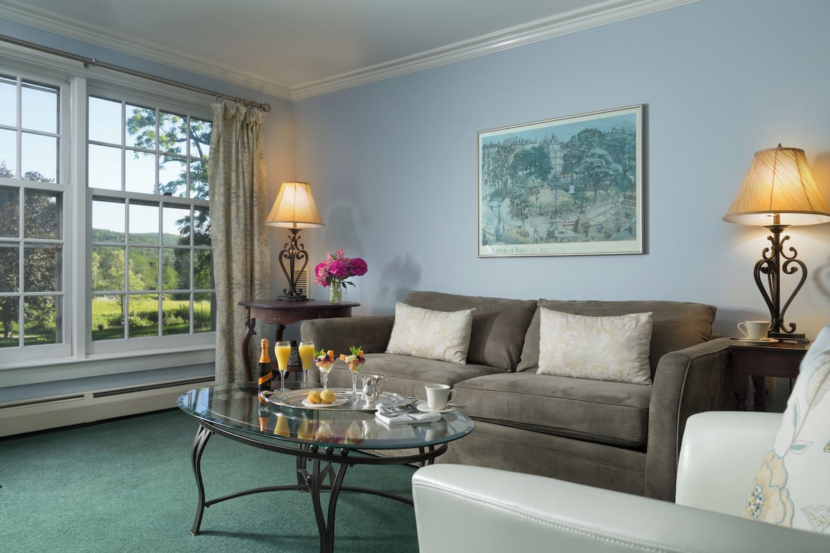 A living room with a glass coffee table, a sofa, and two lamps. A painting is on the wall, and a window offers a view of greenery.