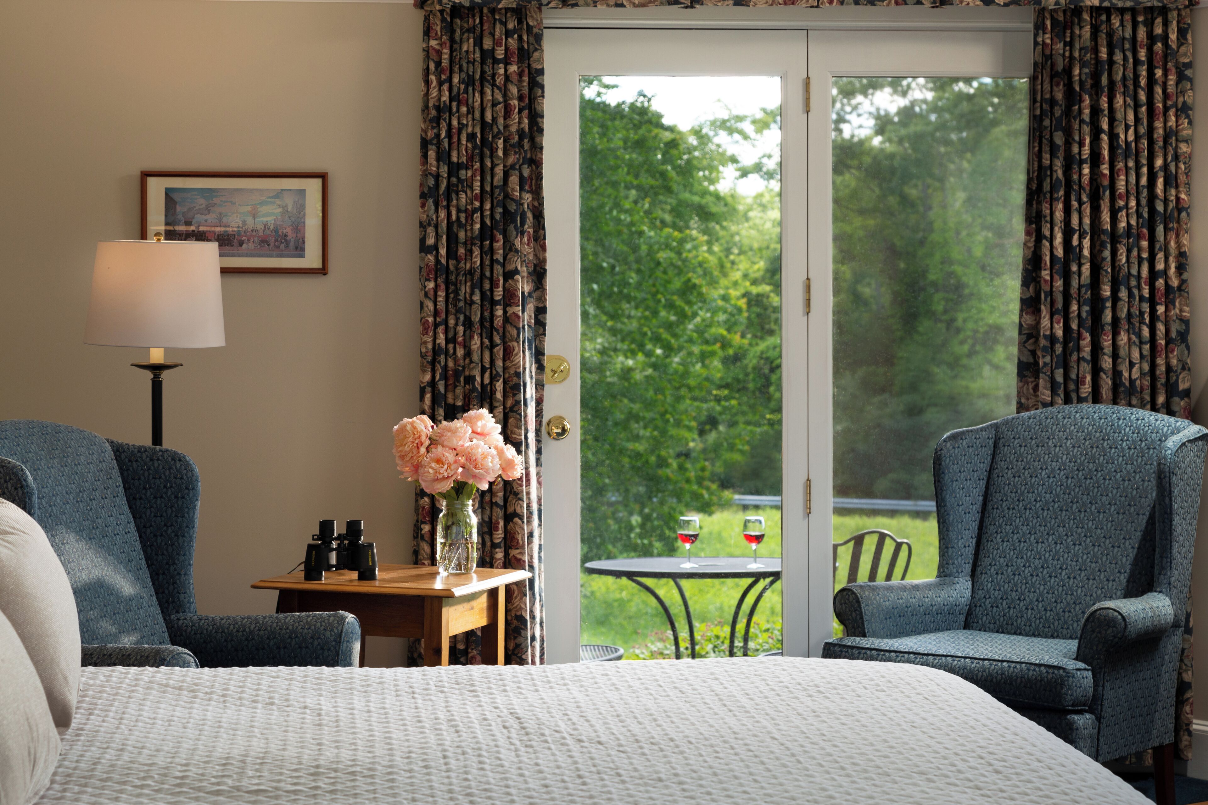 A hotel room with a bed, two armchairs, a small table with a vase of flowers, and a view of a garden through a glass door.