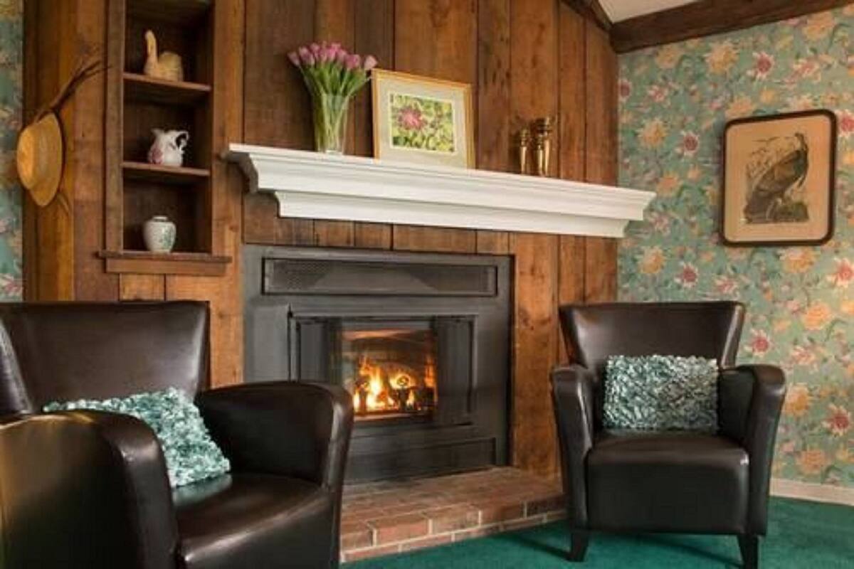 A cozy room with a fireplace, two leather armchairs, a wooden shelf with decorative items, and a floral wallpaper.