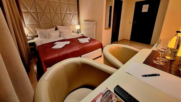 A hotel room with a bed, two chairs, a table with a remote control, a bottle, and a magazine.