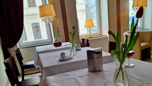 A hotel room with two beds, a dining table with a vase of flowers, a coffee pot, and a wine glass.