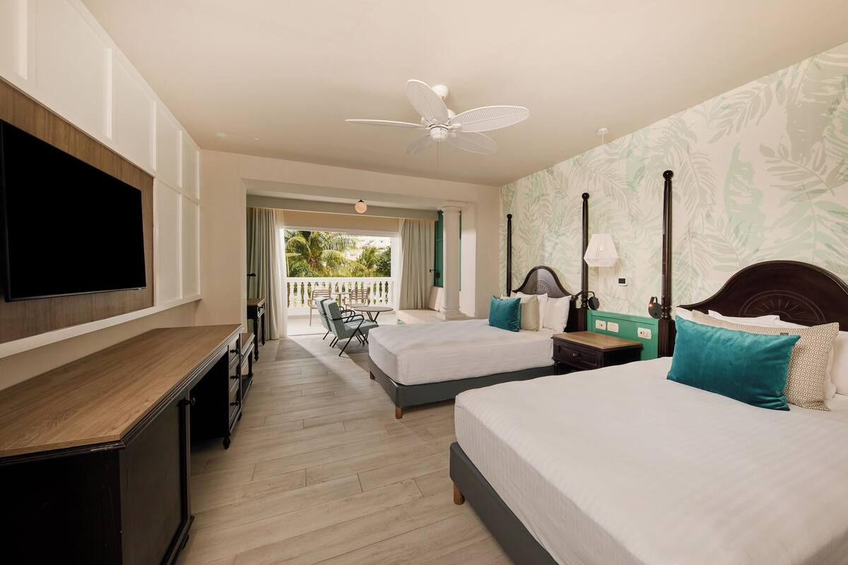 A hotel room with a TV, two beds, a desk, a ceiling fan, and a view of the outdoors.