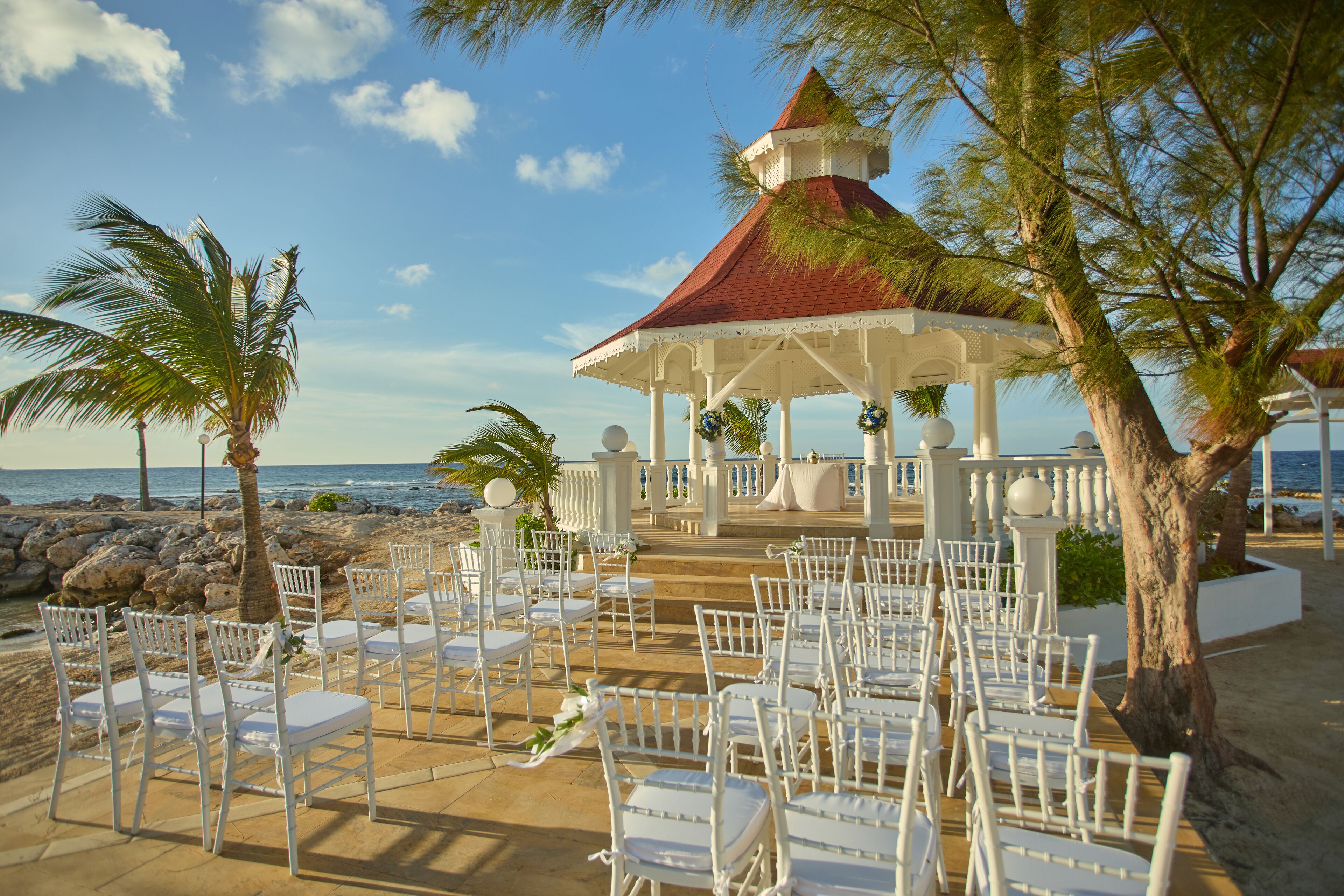 Montaje para una boda frente al mar con sillas blancas y un pabellón.